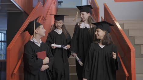 happy caucasian preschool male student in cap and gown holding book and looking at the camera while his classmates going down the stairs at the graduation ceremony