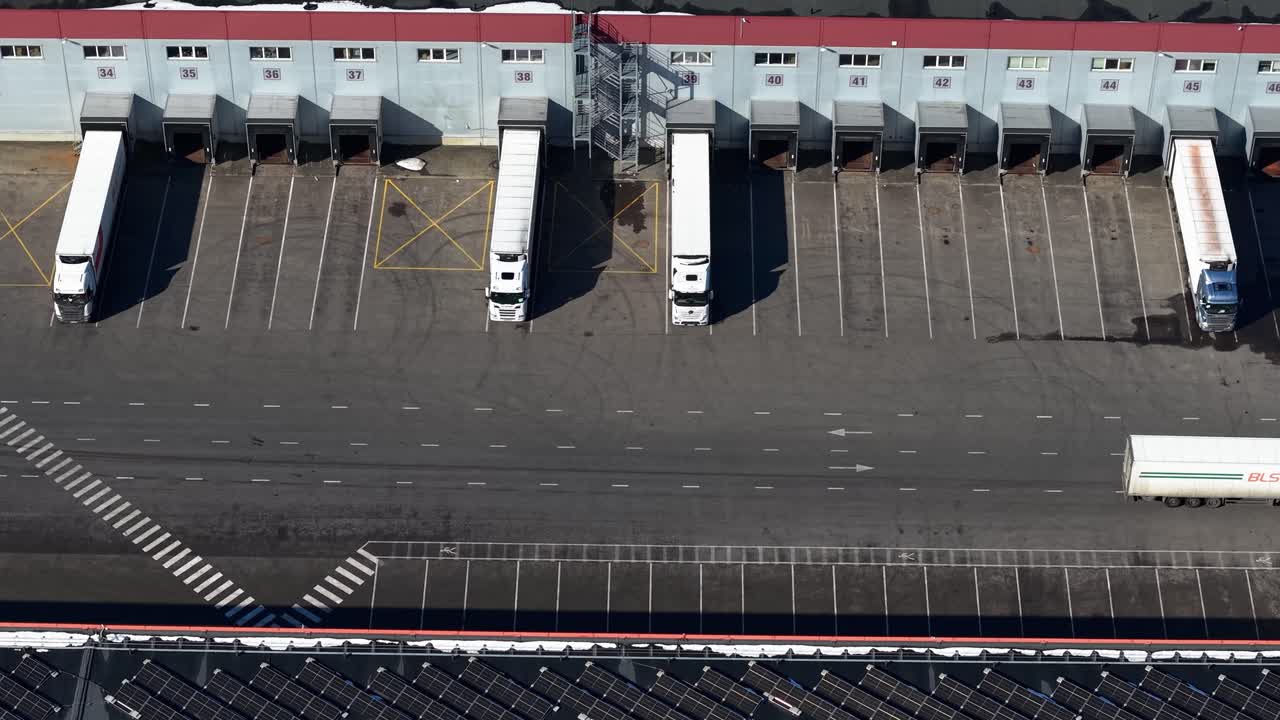 Aerial footage showcasing a busy warehouse logistics hub with multiple trucks at loading bays. The expansive layout, strategically parked vehicles, and clear sky highlight efficient operations