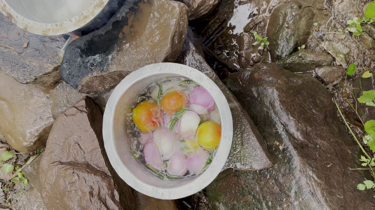 A metal bowl with onions, tomatoes, and herbs being washed at natural stream water among wet rocks during the picnic