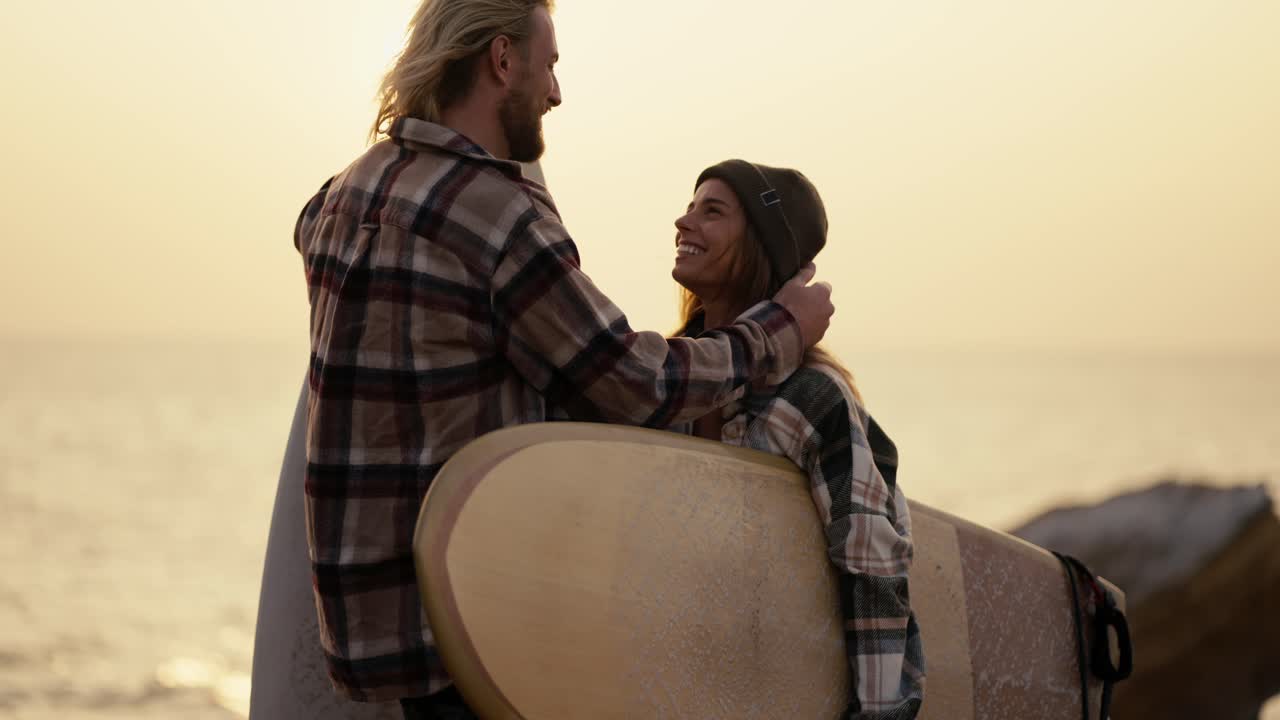 A blond guy in a plaid shirt with a beard is having fun and rejoices while standing and talking with his blond girlfriend in a plaid shirt who is holding a surfboard on a rocky shore near the sea in the morning