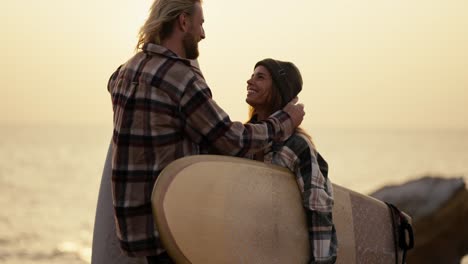 un chico rubio en una camisa a cuadros con una barba se está divirtiendo y regocijando mientras está de pie y hablando con su novia rubia en una camiseta a cuadros que está sosteniendo una tabla de surf en una orilla rocosa cerca del mar por la mañana