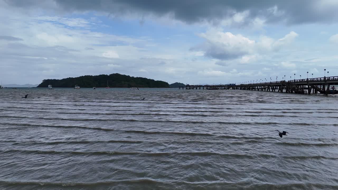 A tranquil aerial view of a pier in Phuket, Thailand, with birds flying over calm waters under a cloudy sky