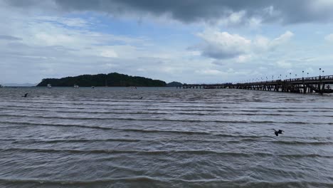 a tranquil aerial view of a pier in phuket, thailand, with birds flying over calm waters under a cloudy sky