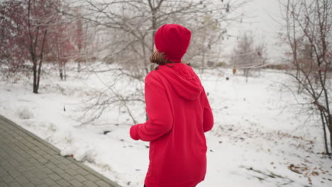 lady wearing red hoodie and beanie jogging along paved snow-covered path outdoors during winter, surrounded by barren trees and serene park scenery, hair swaying in motion