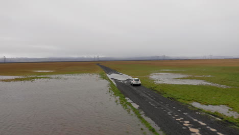 aerial: following a white car on muddy dirt road, driving through a puddle of water