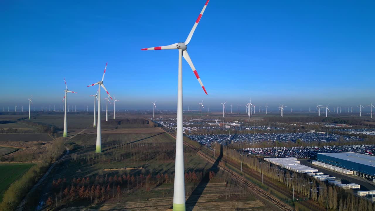 Wind turbines spinning, producing clean renewable energy in a large wind farm, industrial facilities and parking lot below. Unique aerial view ascending raise up drone