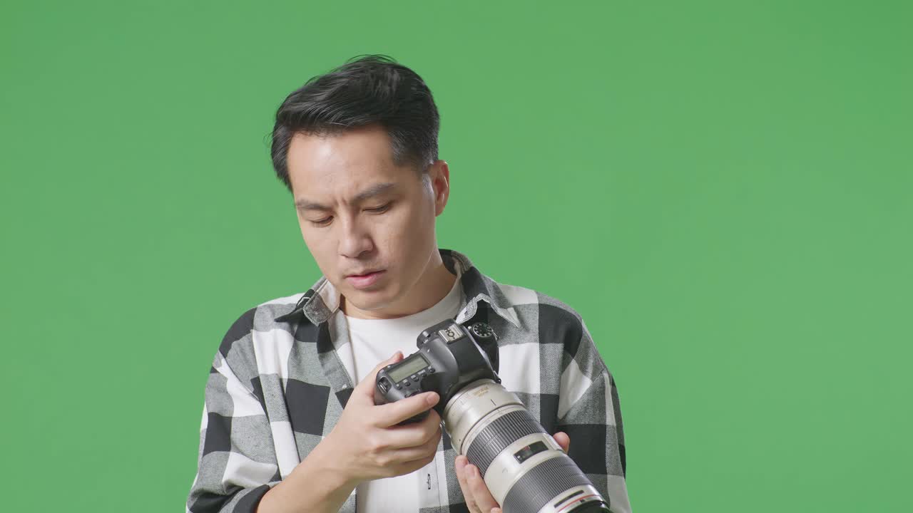 카메라에서 사진을 보는 아시아 사진작가의 클로즈업은 스튜디오에서 녹색 스크린 배경에 서있는 동안 두통을 겪습니다.