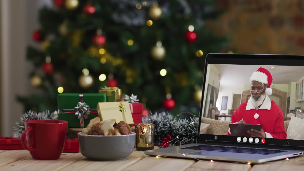 hombre afroamericano senior en disfraz de santa en videollamada en computadora portátil, con árbol de navidad