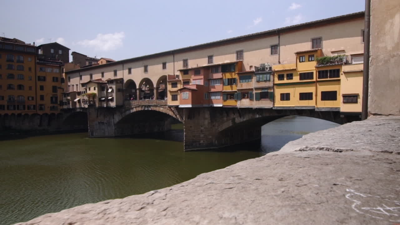 Ponte Vecchio Bridge in Florence, Italy. Tracking towards shot on Gimbal from river bank. Parallax effect with stone wall in foreground.