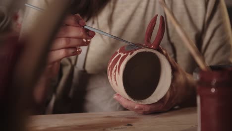 young woman potter drawing a design on the ceramic mug with a brush in art studio