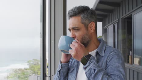 Biracial-man-drinking-coffee-and-talking-on-smartphone-on-balcony-alone