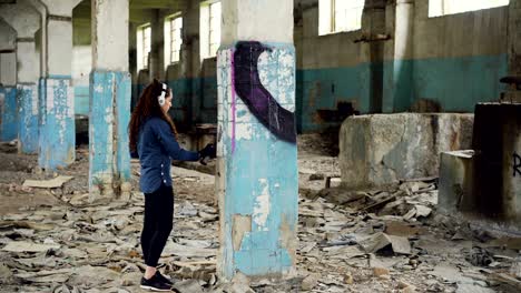 urban artist is decorating column in abandoned warehouse with abstract image using aerosol paint. girl is wearing casual clothing and listening to music with headphones.