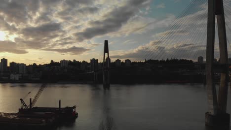 Sky-bridge-suspension-train-track-spanning-over-river-metal-wire-cables-connected-to-cement-towers-Aerial-ascending-wide-angle-blue-sky-clouds-city-bridge-in-background-panning-left-revealing-sunset