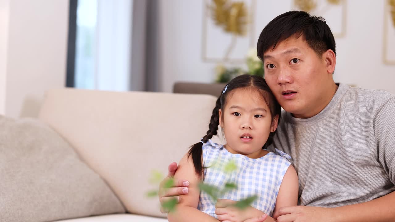 Asian father and daughter react emotionally to suspenseful movie on cozy living room sofa
