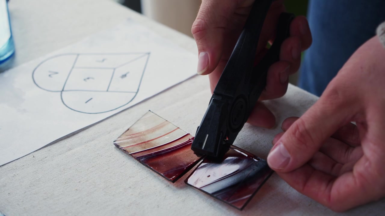 A male art student successfully scores and cuts a small piece of swirled orange glass during a stained glass class. His design template is visible in frame.