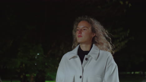 focused girl in white coat walking outdoors during evening stroll, ready to sit and read book, soft streetlights glowing, dark background with three girls passing by