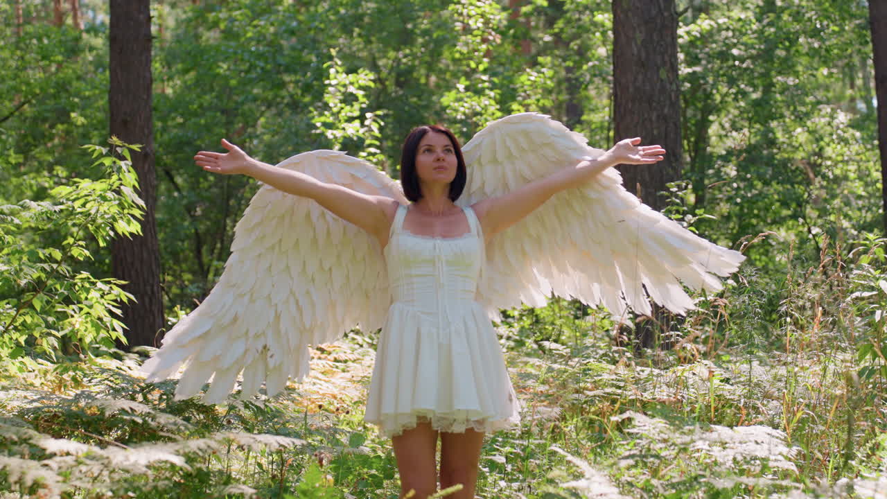 Fairy white woman with large angel wings walking gracefully through bright woodland lifting arms toward sunlight, surrounded by glowing leaves and warm forest greenery