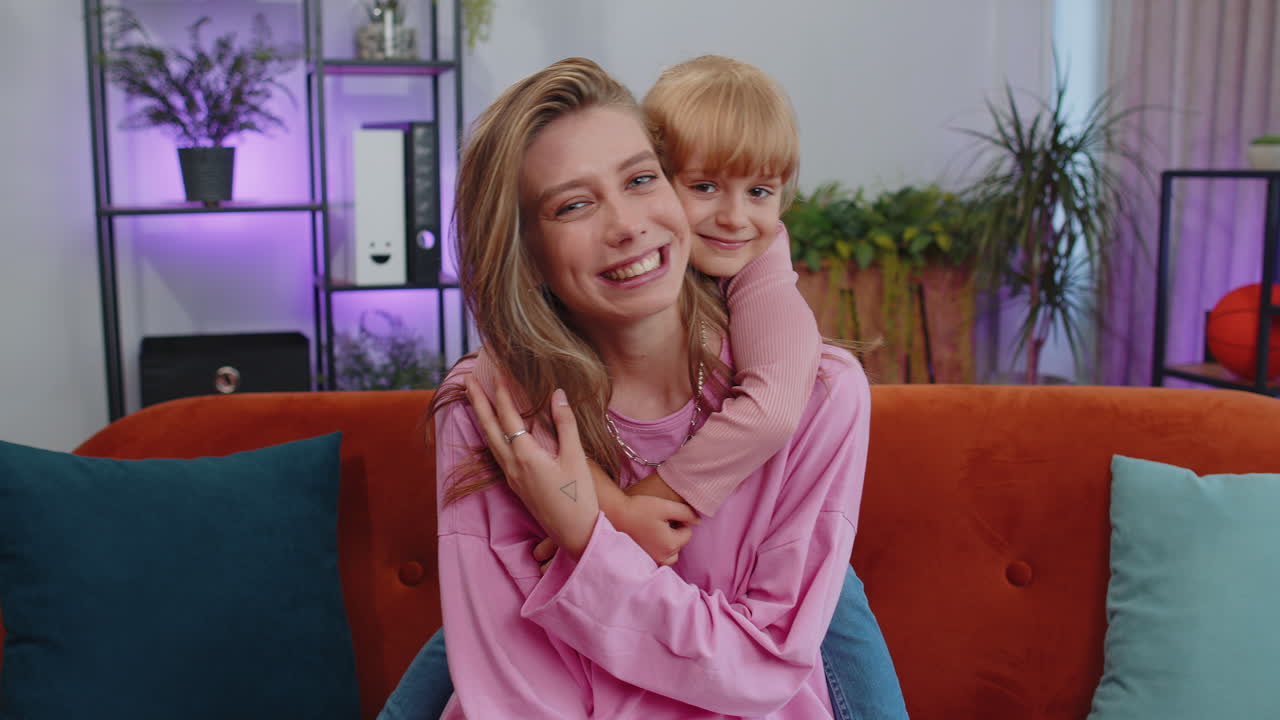Smiling child kid daughter embrace young mother, look at camera, sweet tender love at home sofa