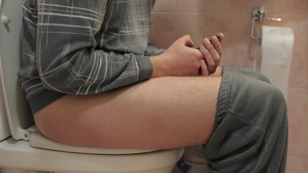 Man Sitting On Toilet Use Cell Smart Phone Young Guy Chatting Online In Restroom
