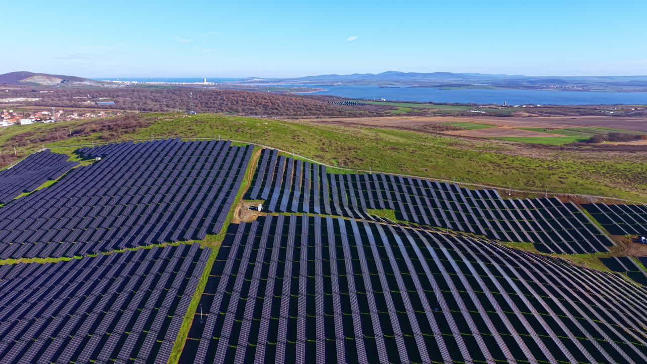 Stor solcellepark på en skråning nær en sø med grønne marker og fjerne bjerge på en klar dag
