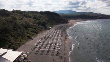 Drone pushes over Ahtopol beach, capturing sun umbrellas, loungers, bars, people relaxing, and waves crashing along the shore