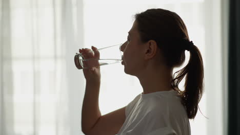 woman drinks clear water standing by window. pretty lady enjoys refreshing cold liquid from glass at home slow motion. morning healthy routine