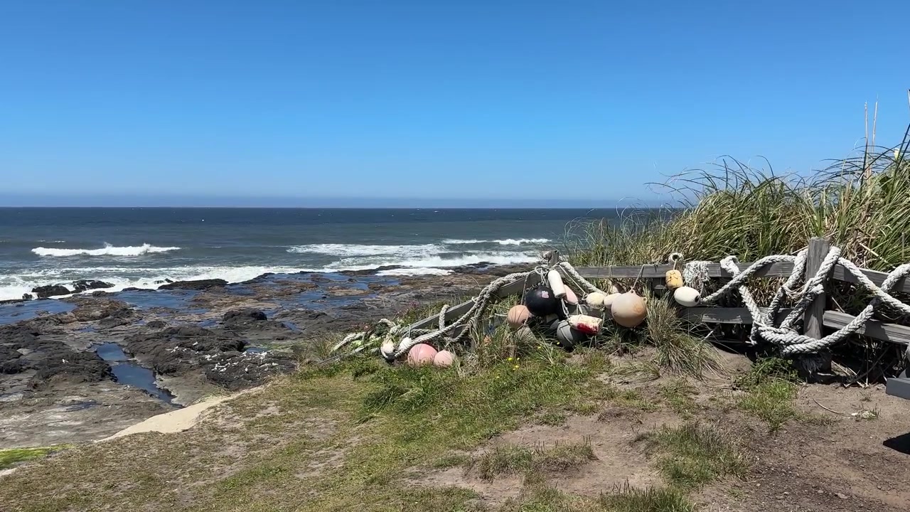 Boje i pływaki zawieszone na linach wiszą na zwietrzałym płocie nad skalistym wybrzeżem Oregonu, z basenami pływowymi i rozbijającymi się falami wzdłuż wybrzeża Pacyfiku.