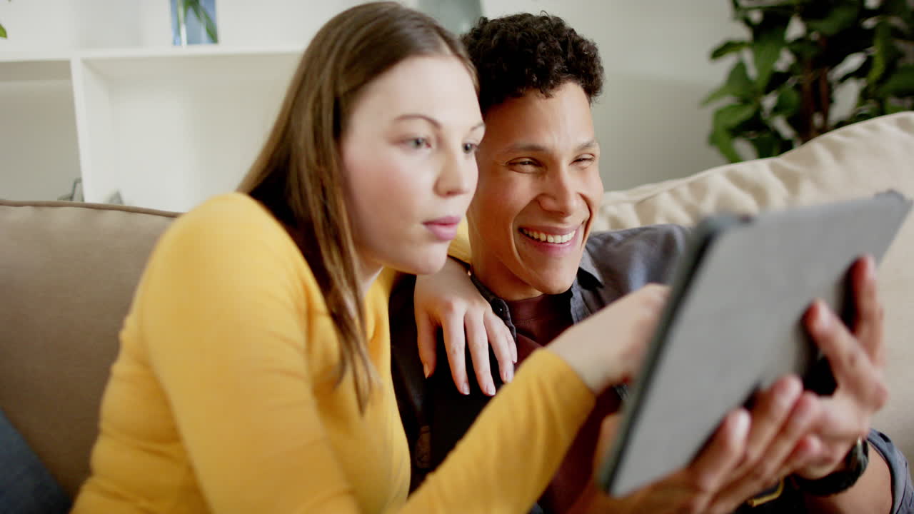 Casal feliz e diverso sentado no sofá usando tablet em casa, em câmera lenta