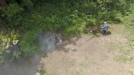 aerial view of a man operating a tiller near a smoking debris pile in a natural, overgrown area