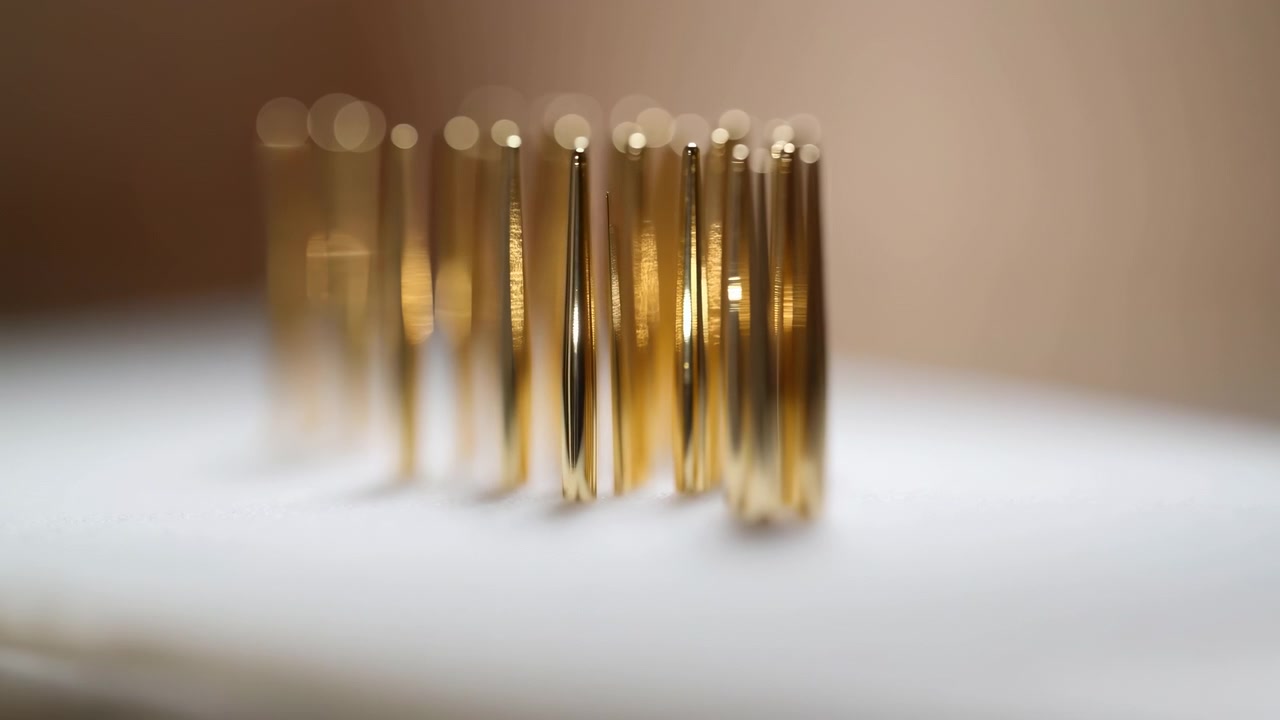 Perni cilindrici dorati stanno in piedi su una superficie bianca, con l'area centrale nitida mentre le file esterne si sfumano in un effetto bokeh, creando un elegante motivo commerciale astratto con caldi riflessi luminosi.