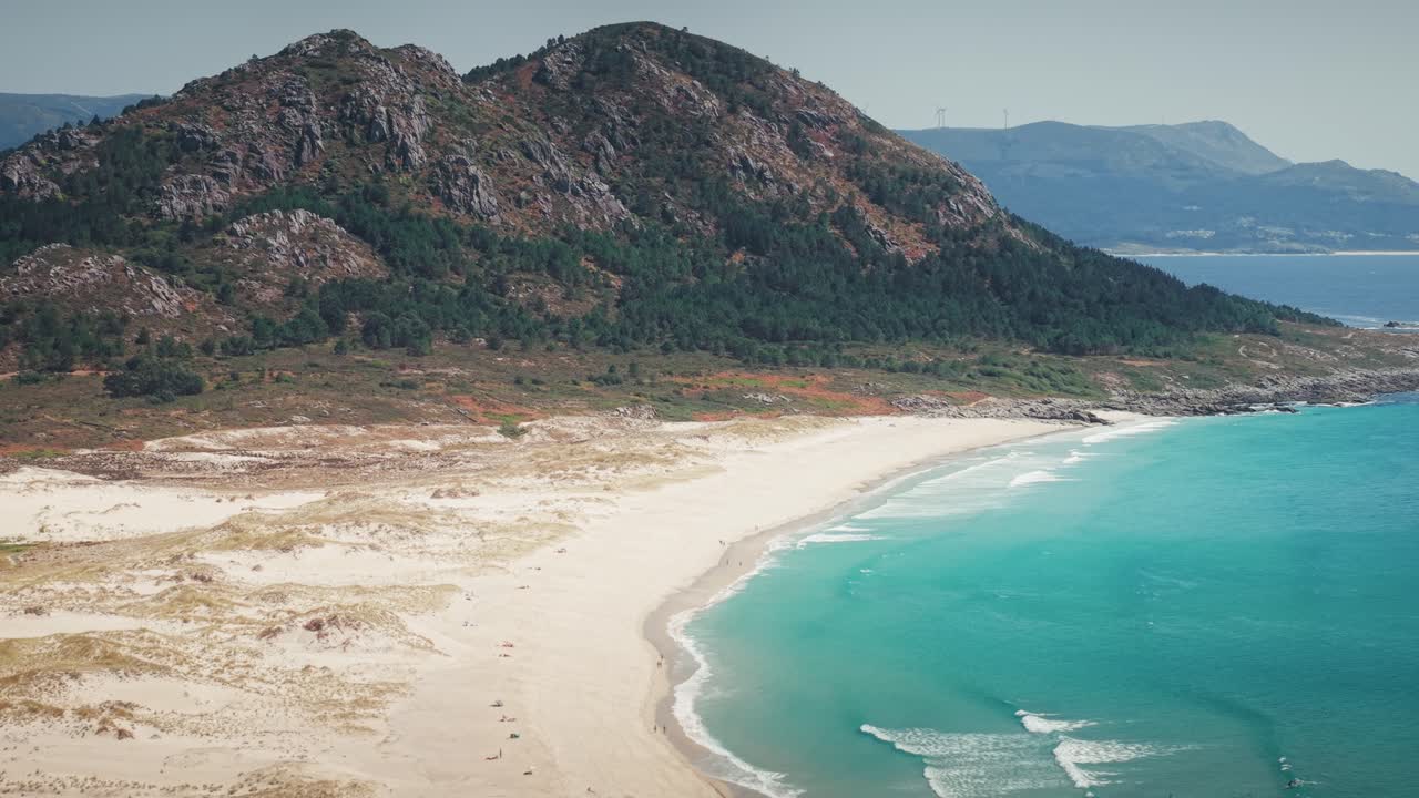Ripresa laterale con teleobiettivo da drone lungo la spiaggia di Monte Louro in Galizia, Spagna, con acqua turchese e sfondo montuoso.