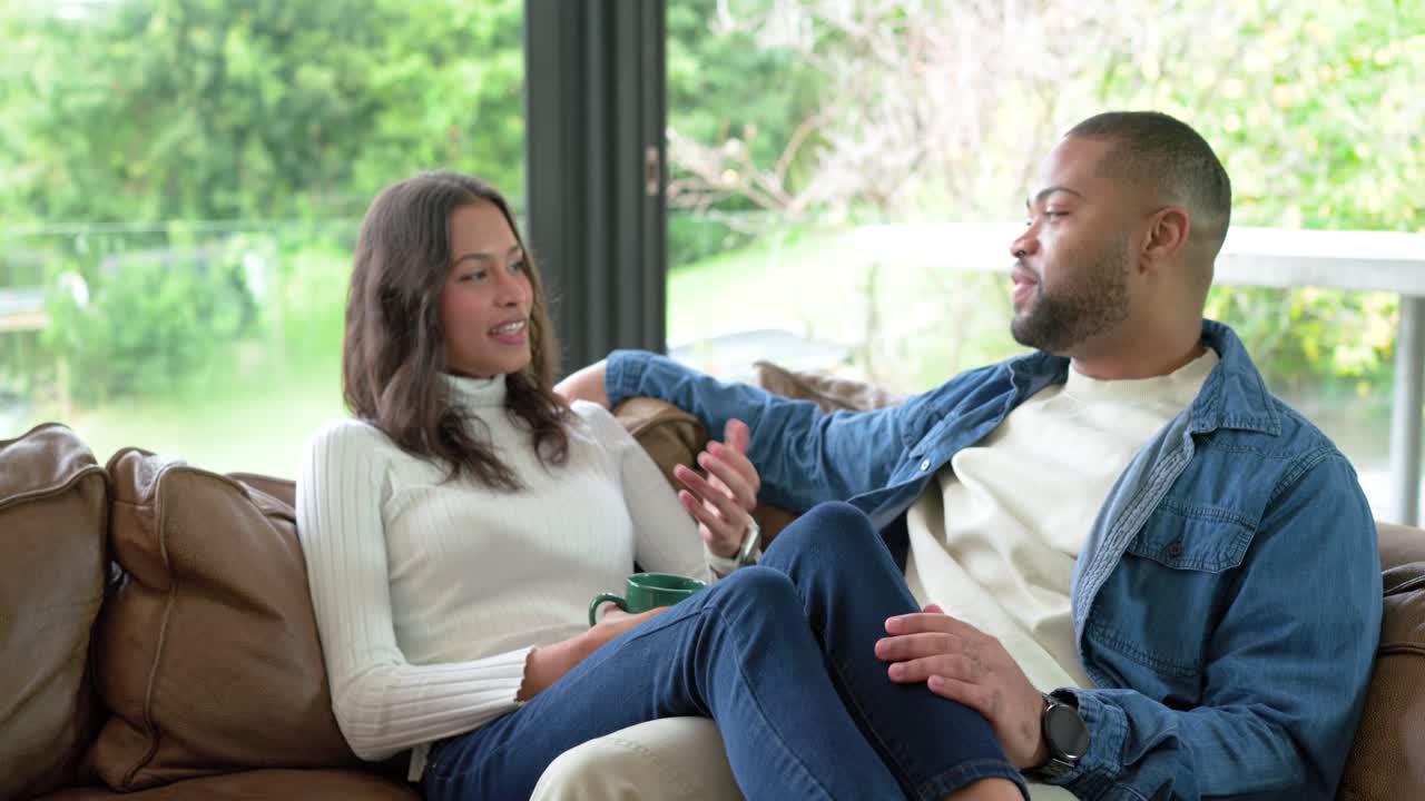 Diverse couple settling onto leather sofa by window, talking and laughing over green ceramic mug