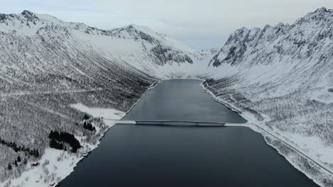 Drone-view-in-Tromso-area-in-winter-flying-over-a-fjord-surrounded-by-white-mountains-and-a-bridge-crossing-in-Norway