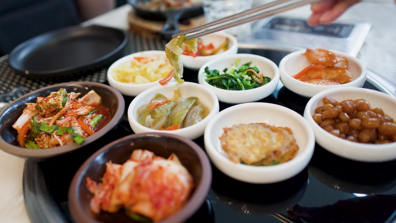 금속 젓가락이 작은 흰색 세라믹 그릇에서 다양한 발효 채소와 반찬을 집어 올린다.