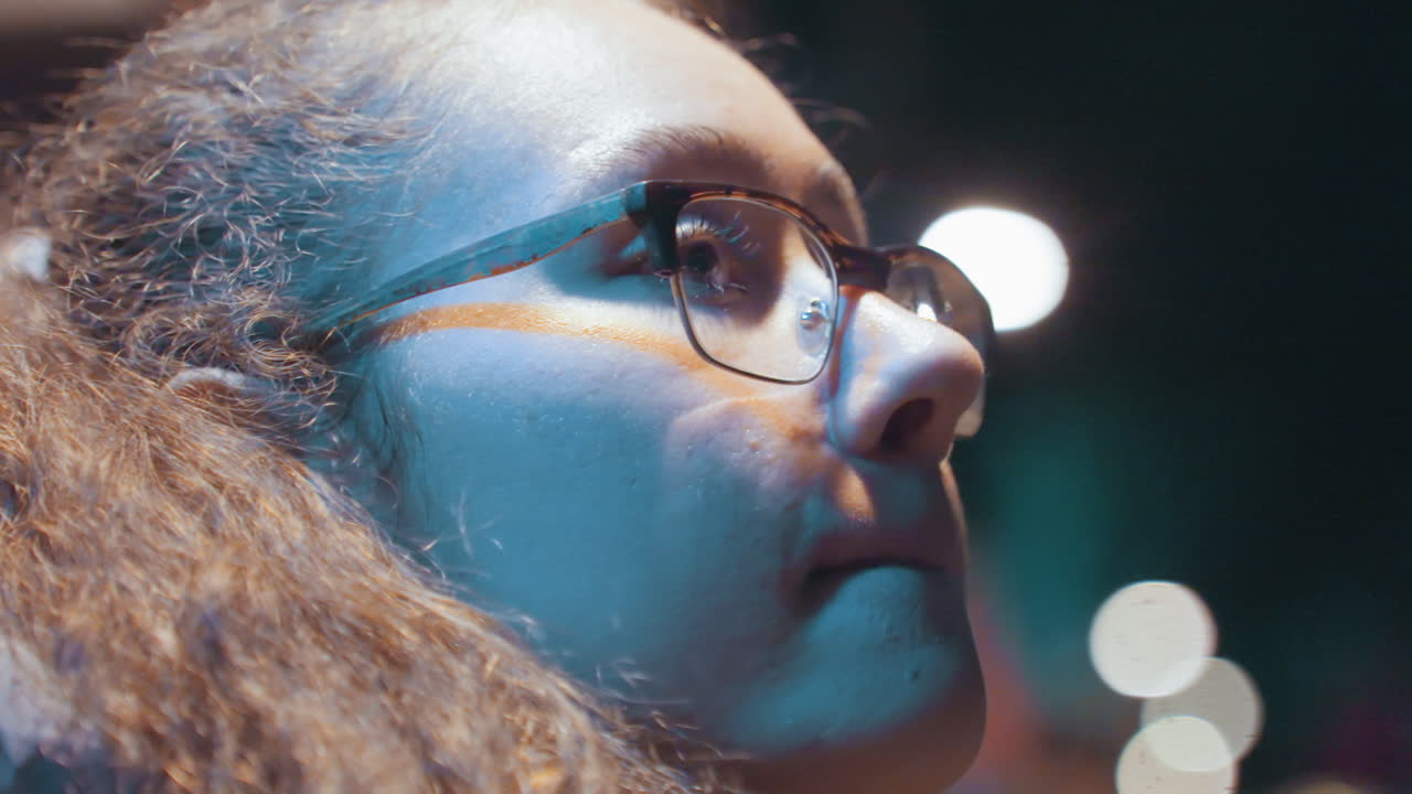 Nighttime street close up woman with glasses, curly hair, bokeh lights, neon glow latenight snack moment, contemplative gaze, slow breath, subtle film grain, candid festival or cinema queue vibe