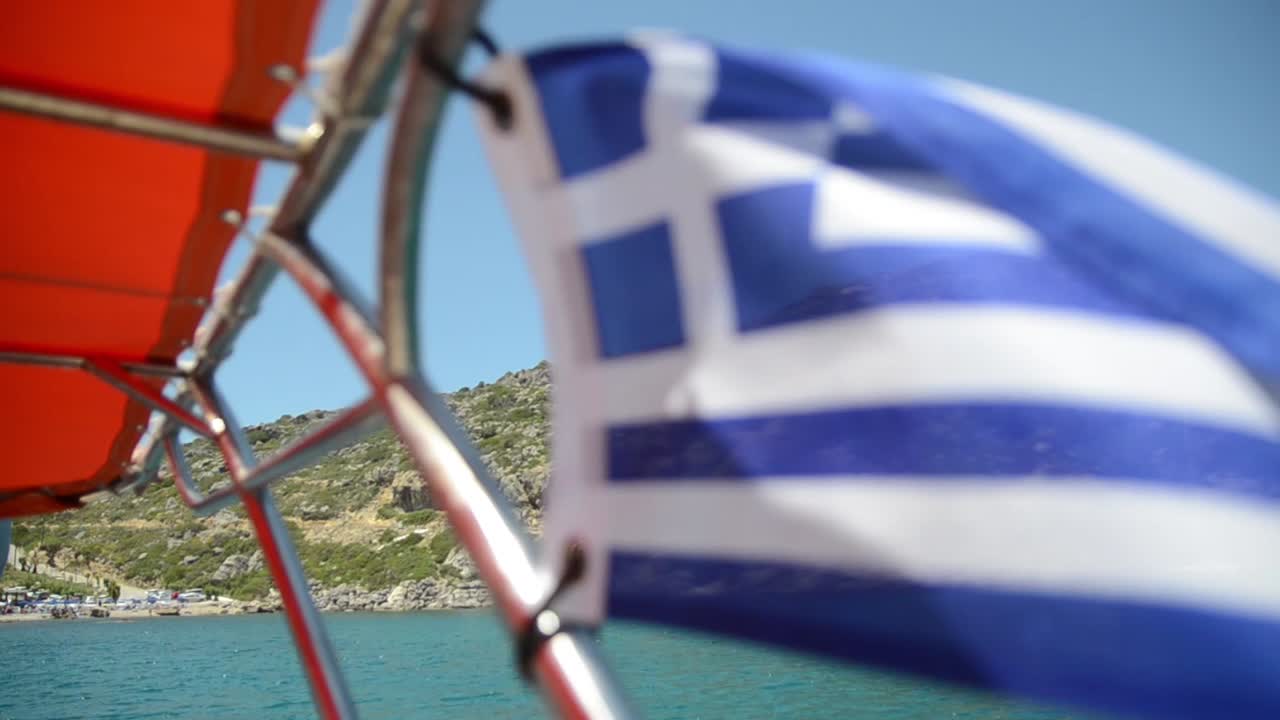 Flag of Greece flying in the wind on boat on sea, focused on island on background