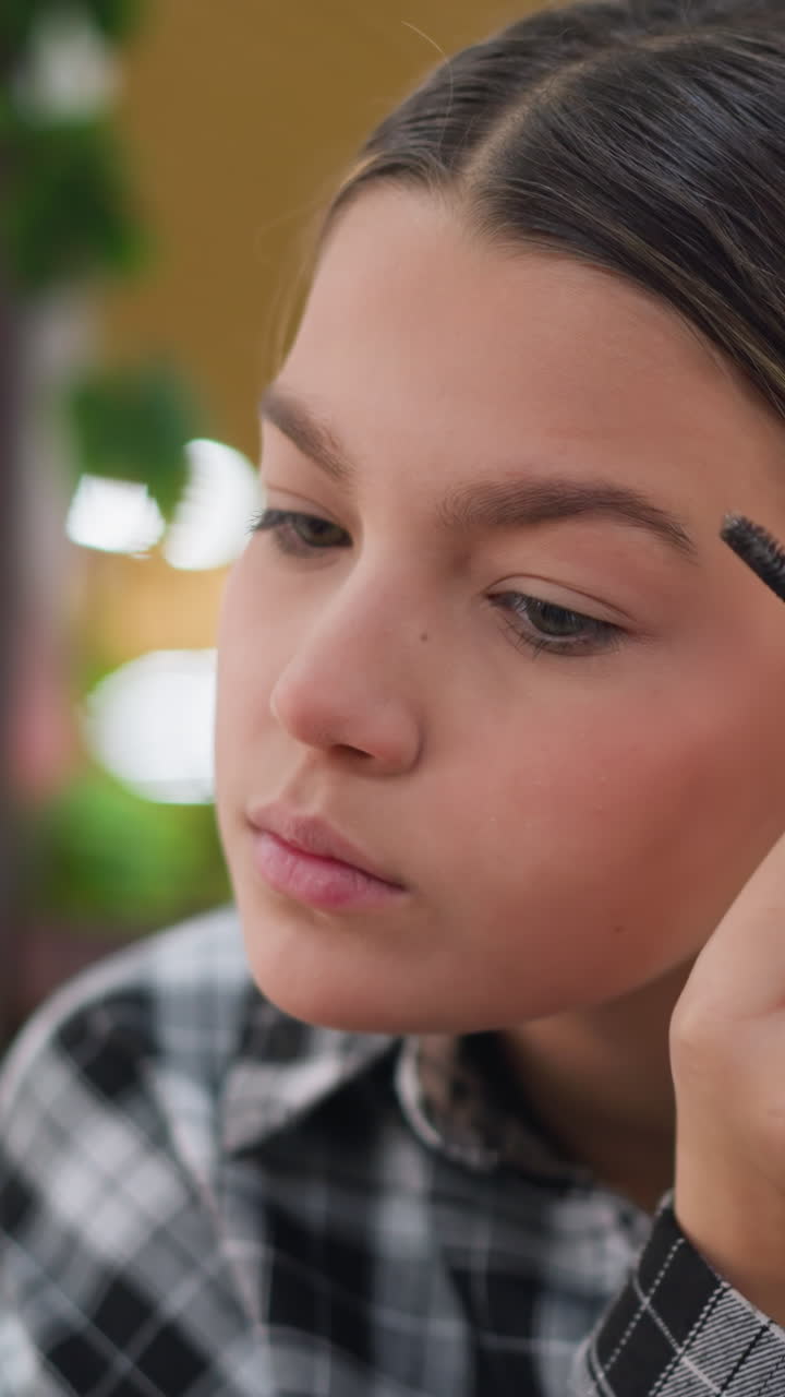 Adolescente caucásica dándose forma a las cejas con un cepillo en la mesa de una cafetería, camisa de cuadros, fondo bokeh suave, expresión concentrada, ambiente de creación de contenido, cuidado preciso, práctica de tutoriales, día informal