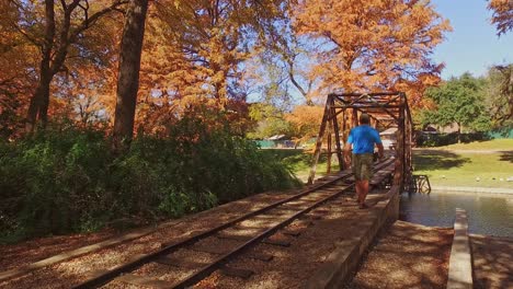 Hombre-Caminando-Por-Las-Vías-Del-Tren-En-Miniatura-Hasta-El-Puente-En-Miniatura-En-El-Parque-Hermosos-Colores-Otoñales-De-Los-árboles-Del-Parque-Alrededor