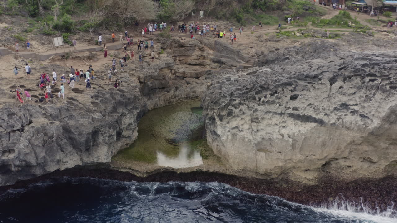 Small lagoon trench in the coast of Broken Beach, with tourists taking photographs on its sides. Nusa Penida, Bali, Indonesia. Aerial shot