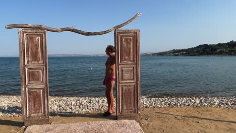 Ungewöhnliche-Aufnahmen-Einer-Holztür-Am-Strand-Für-Dreharbeiten-Mit-Blick-Aufs-Meer,-Mit-Einem-Mann-In-Badebekleidung,-Der-Sich-An-Den-Türrahmen-Lehnt