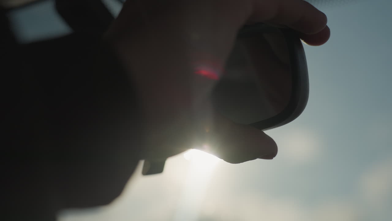 person adjusting driver mirror sunlight streaming through window creating dynamic lens flare inside car cabin urban backdrop as hand adjusts mirror for glare control cinematic lighting