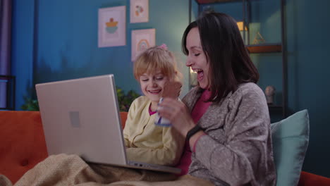 madre y niño pequeño hija niño estudio en la computadora portátil, viendo películas divertidas, relajándose, acostado en el sofá