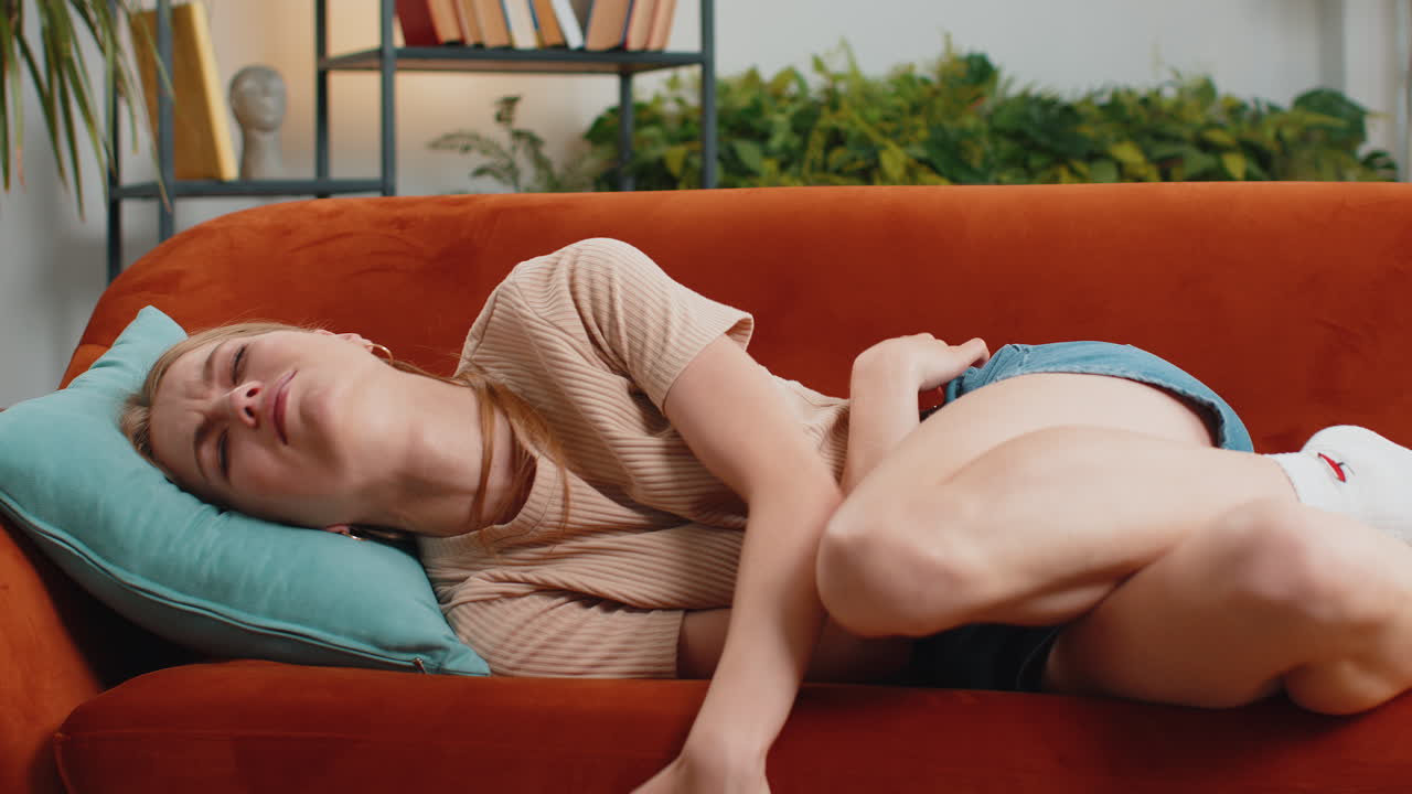 Sick ill woman suffering from period cramps, stomach ache menstrual pain lying on sofa at home room