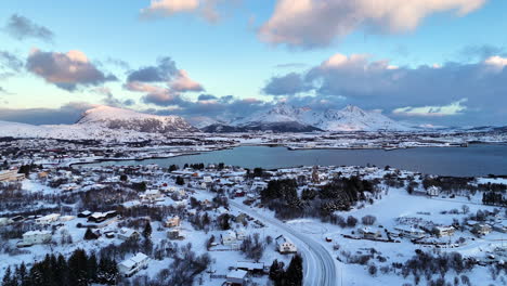 Aerial view of winter town in Lofoten, Norway, surrounded by snow-covered houses, mountains, and dramatic arctic landscape under soft evening light