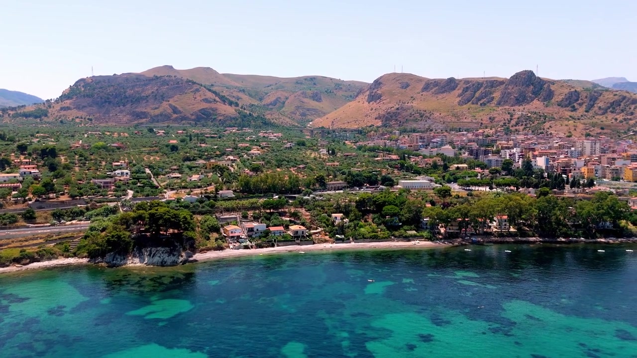 Voo de drone sobre a vibrante costa mediterrânea revelando assentamentos costeiros aninhados sob montanhas escarpadas em Palermo, Itália