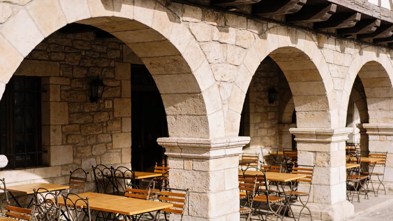Porticato in pietra centrato che reagisce al sole che si sposta sulla terrazza del caffè, con tavoli in legno