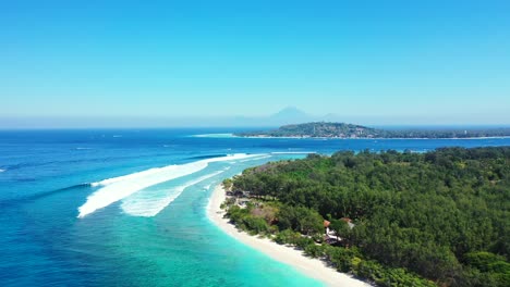 Grandes-Olas-Blancas-Salpicando-En-La-Playa-De-Arena-Blanca-De-Una-Isla-Tropical-Con-Una-Exuberante-Vegetación-Rodeada-Por-Una-Laguna-Azul-Turquesa-En-Bali