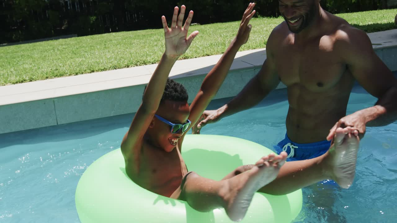 African American father and son nudging neon ring at pool, boy kicking, father steadying for fun