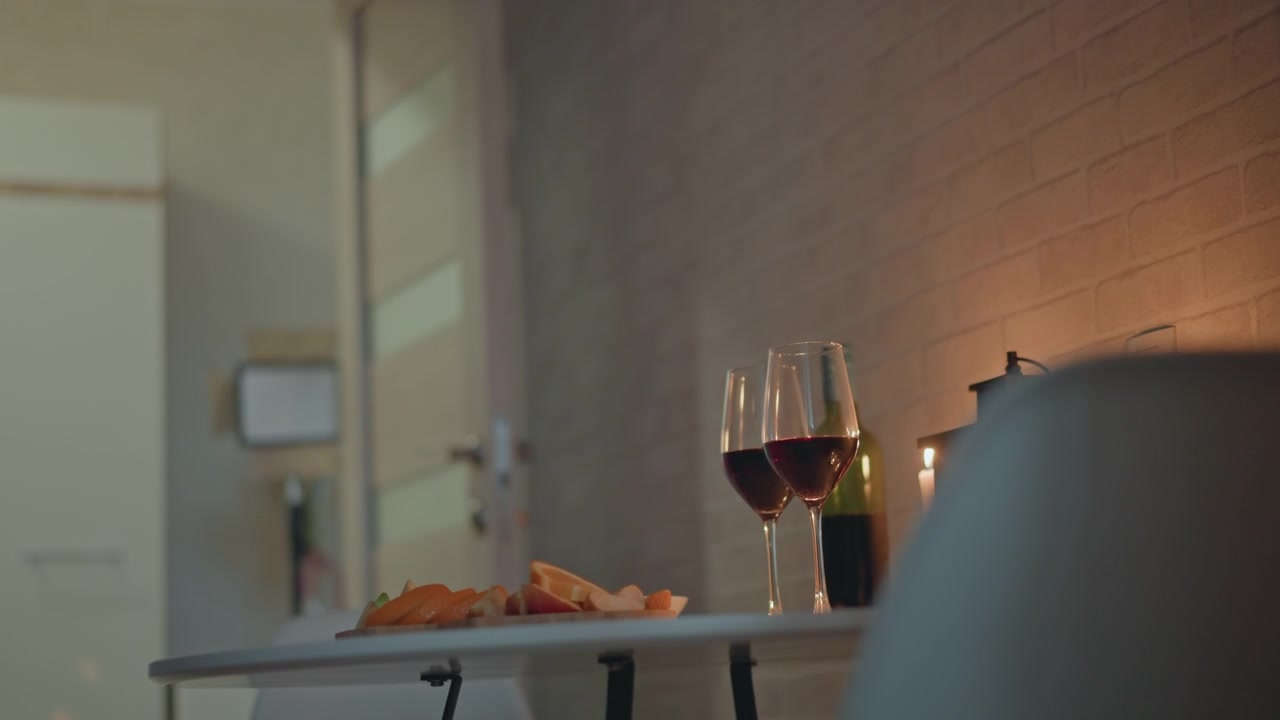 Low-angle interior scene showing two filled red wine glasses beside pastry plate and salt and pepper shakers on compact tabletop, with chair partially obscuring right side and warm wall glow