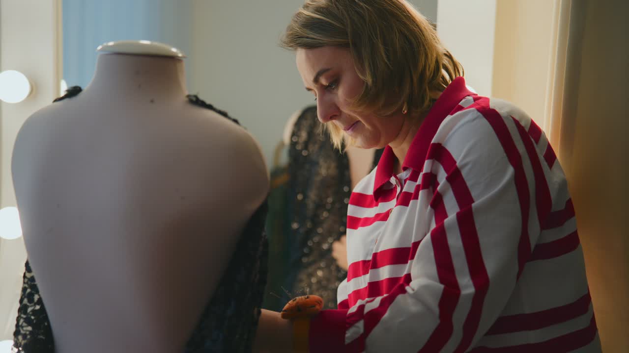 Donna sistema attentamente un capo d'abbigliamento su un manichino in uno studio luminoso. La sua concentrazione è intensa mentre liscia il tessuto. Indossa una camicia a righe rosse e bianche. Design di moda o prova di abiti.